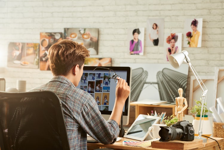 Graphic designer reviewing portfolio PDF on computer in creative workspace with photos and camera.