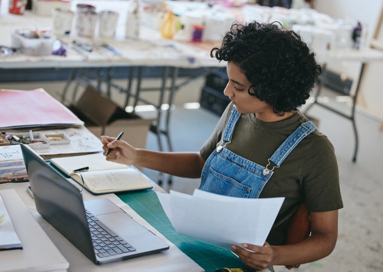 A female graphic designer in a creative studio in India, working on a laptop and taking notes about a project.