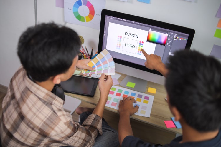 Graphic designers in a creative workspace evaluating logo designs and colour choices on a laptop screen and comparing them with on-hand colour swatches.