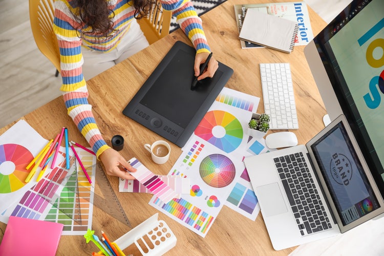 Designer working on brand identity design with colour palettes, graphics tablet, and logo on computer screen.