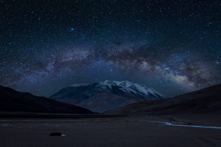 Bow-shaped Milky Way arching above a round mountain peak, with the night sky illuminated by countless stars.