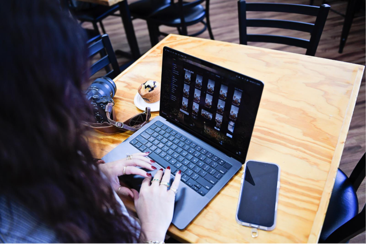 A photographer using Adobe Lightroom to batch edit photos on her laptop at a cafe.