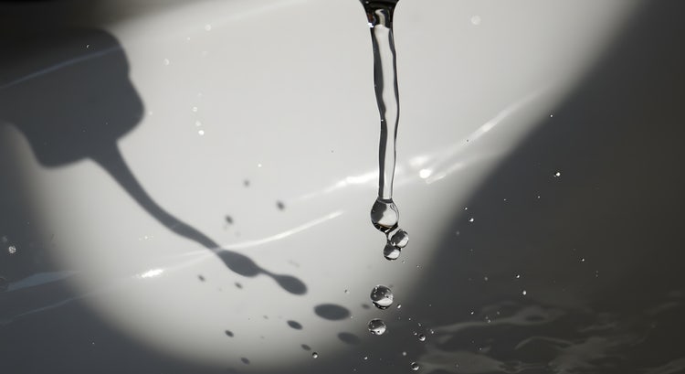 Water droplets falling into a sink with a distinct drop shadow of the faucet and splash visible on the curved surface.