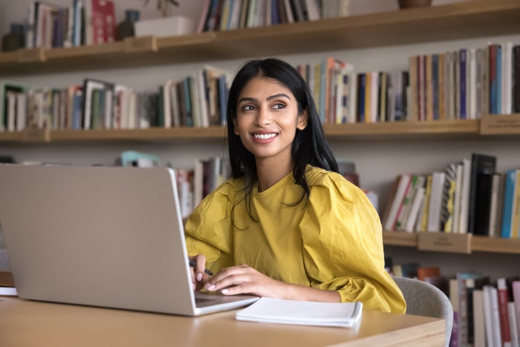 Woman sitting at a desk with a laptop in a library setting, smiling while working through a Photoshop tutorial for beginners.