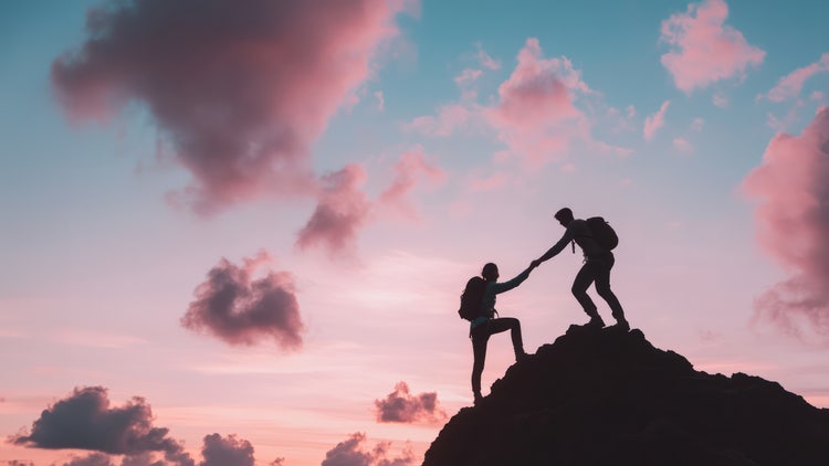 Silhouette photography of two hikers climbing a mountain at sunset, one helping the other, with a warm sky in the background.