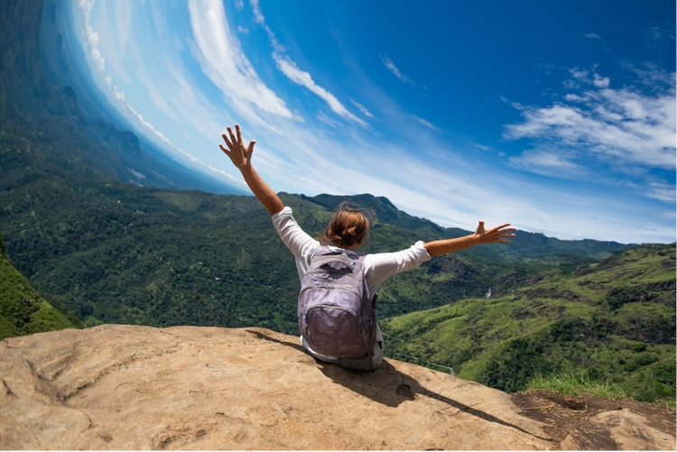 Back view of a woman with a backpack sitting on a mountaintop with arms raised, overlooking a distorted landscape sky caused by rolling shutter distortion.