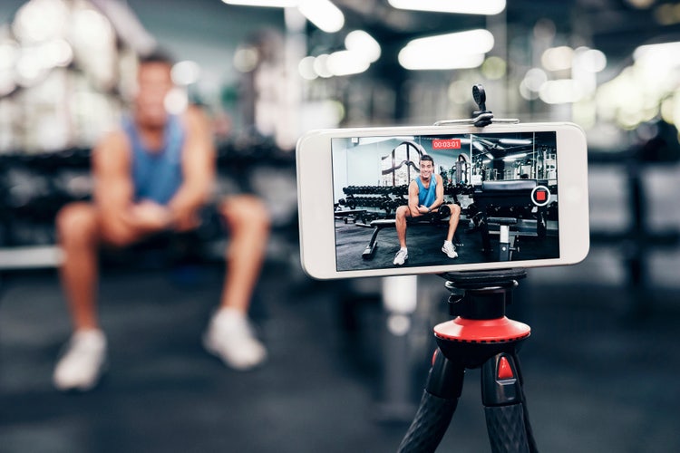 Fitness content creator recording a video on a smartphone in a gym, with focus on the screen showing proper aspect ratio for filming.