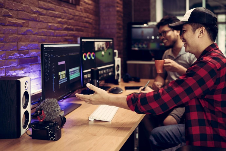 Two video editors review footage on dual monitors in a studio, discussing frame rate settings while editing a project.