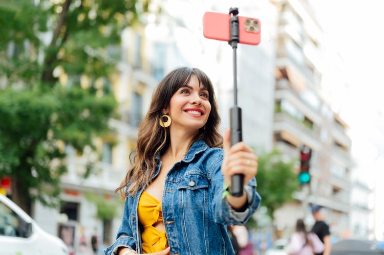 Smiling woman recording herself with a smartphone on a selfie stick in an urban setting, representing creators converting Live Photos to video for vlogs and social media content.
