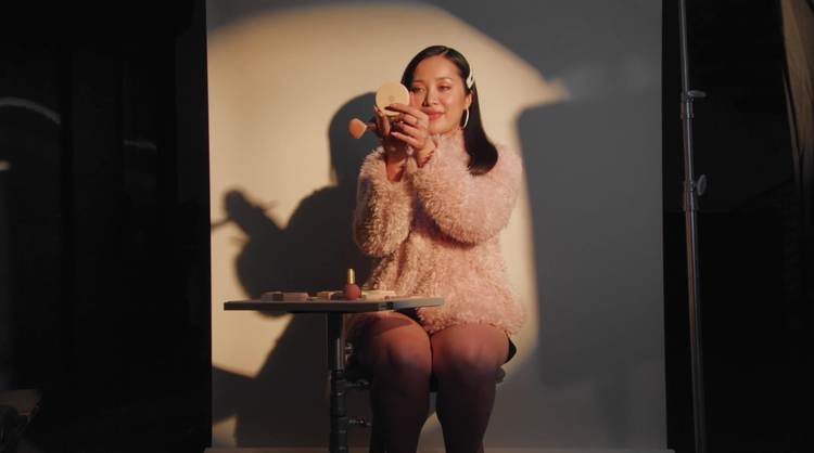 Portrait of creator Michelle Phan in a shaggy pink pullover, holding a makeup compact and brush.
