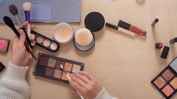 A shot for a faceless video showing a laptop, a pair of hands, and a variety of lipsticks, blushes, and brushes atop a wooden table.
