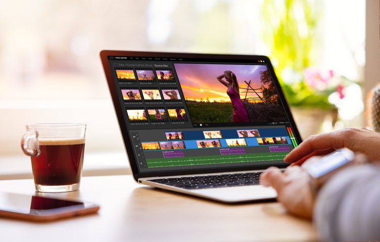 A pair of hands working on a laptop, using a video editing software to edit a footage. A cup of coffee and a table is on the left side.