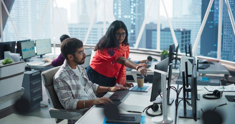 Two Indian professionals collaborating at computer workstations in a modern office with city view