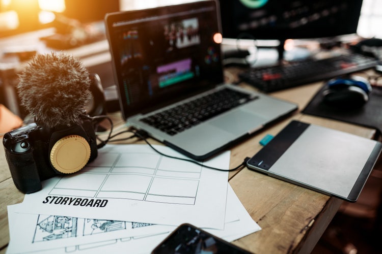 A messy desk containing a laptop, a computer, a camera, a drawing table, multiple storyboard papers, and other creative tools for video creation.