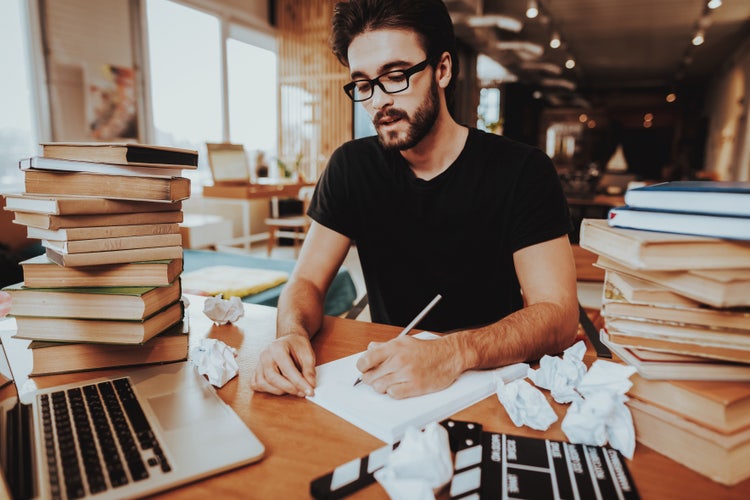 A male writer in working on a script, holding a pen over a stack of paper, surrounded by piles of books, crumpled papers, and a laptop.