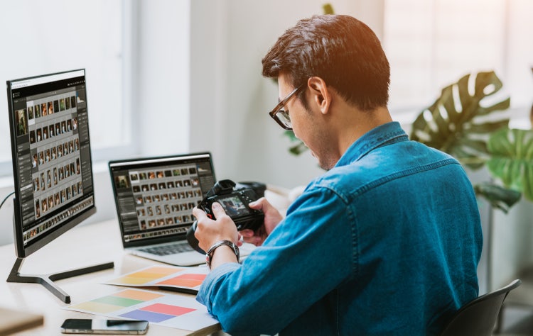 Photographer using object remover software on computer to edit photos, with camera and color swatches on desk.