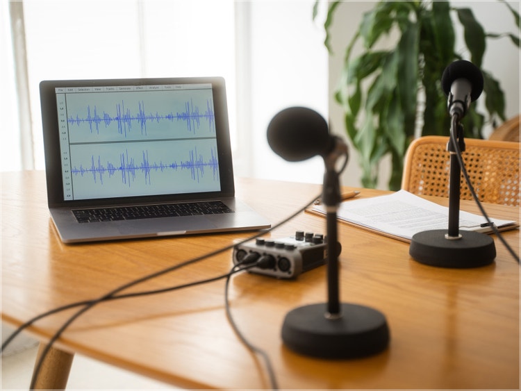 Laptop with audio waveform, microphones, and mixer on wooden desk for a recording session.
