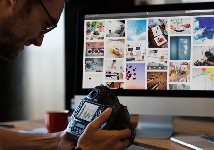 Photographer reviewing images on digital camera beside computer displaying AI image style transfer gallery.