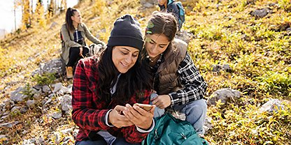 Two people sitting next to each other outside and looking at one of their phones together
