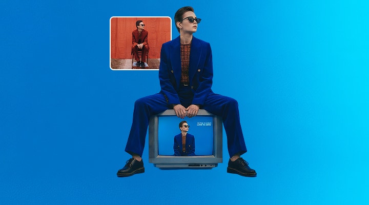 A woman in an orange suit sits on a chair in a field. The Photoshop AI Assistant is used to swap in a blue background, to replace the chair with TV and change the suit's colour to blue.