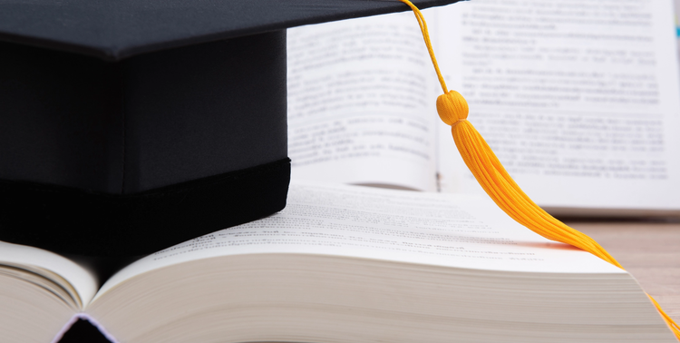 Cappello di laurea con nappa gialla appoggiato su un libro accademico con un altro volume sullo sfondo.