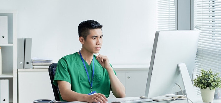 Un uomo con un camice verde seduto alla scrivania, davanti a un computer.
