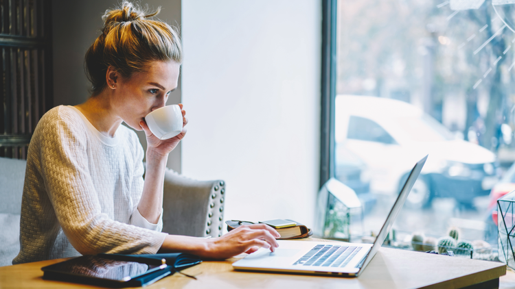 A woman with blonde hair sips out of a mug while browsing on a laptop.