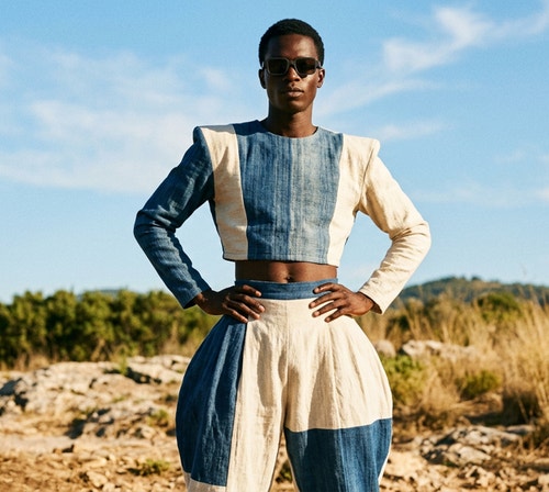 Persona con occhiali da sole e un outfit strutturato bianco e blu in piedi all’aperto, sullo sfondo di un cielo terso e un paesaggio naturale