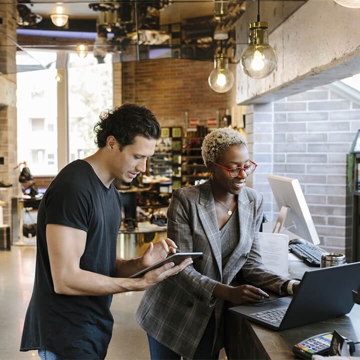 Woman in business attire and a man in a black shirt reviewing business files on laptop and tablet