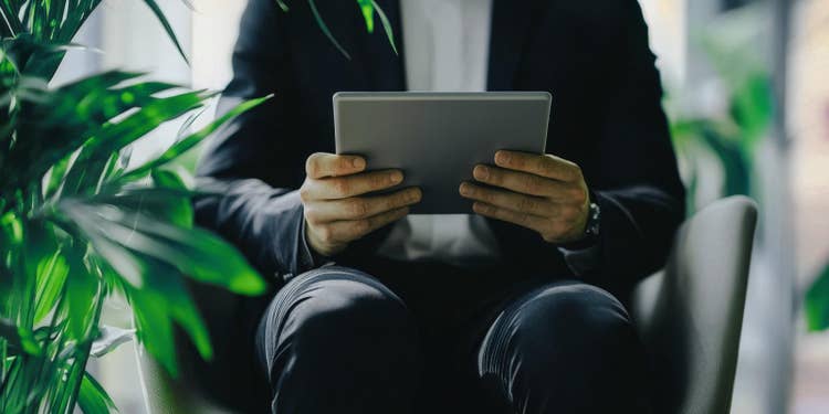 アイボリーの背景にグレーのスーツでタブレットを持ったビジネスマンと観葉植物