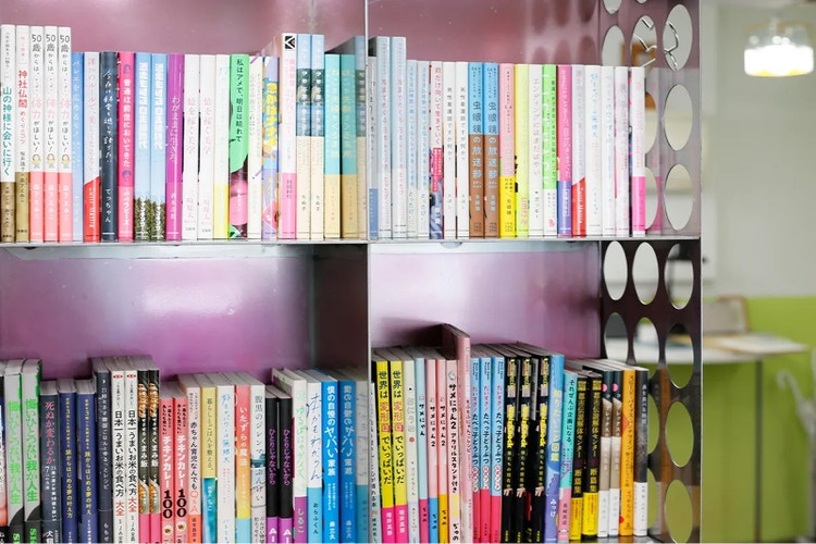 本棚に並んだ多数の日本語の書籍を写した写真。ピンク色の棚にカラフルな背表紙の本が整然と並び、右側には丸い穴が並ぶ仕切りと、ぼかした室内空間が見える。