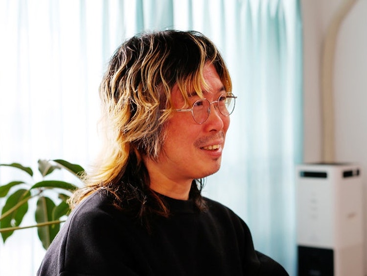 横向きに座る人物のポートレート。黒とブロンドのツートーンヘアにメガネをかけ、黒いトップスを着た人物が室内で微笑んでいる。背景には観葉植物と白いカーテンが見える。