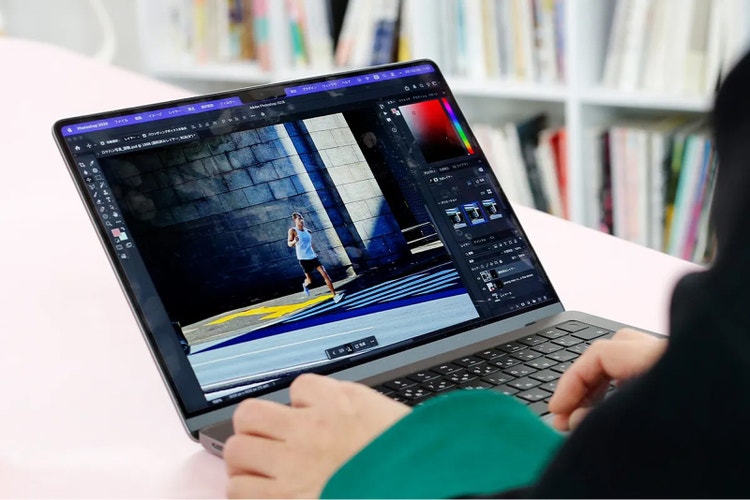 ノートパソコンでAdobe Photoshopを操作している様子。画面には石壁の前を走る人物の写真が表示され、右側にレイヤーやカラー調整の編集パネルが開かれている。背景にはぼかした本棚が見える。