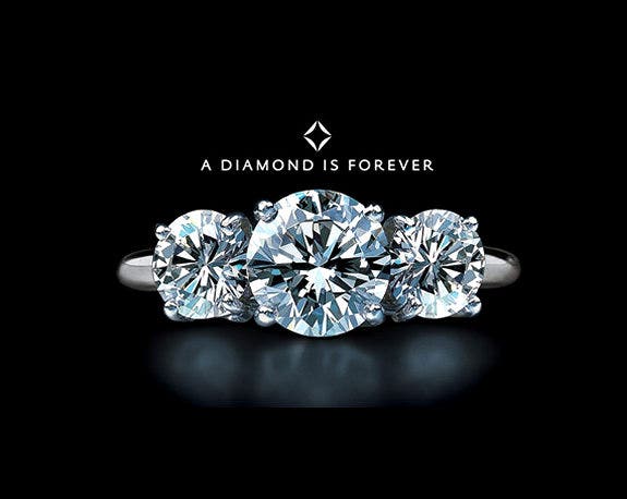 A diamond ring against a black background with the De Beers logo and slogan above