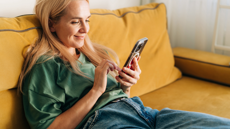 A woman with blonde hair sits on a yellow sofa as she smiles and looks at her phone.