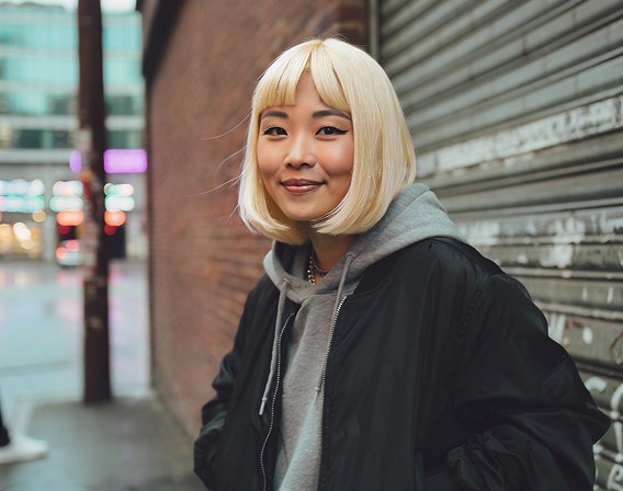 街の通りでレンガの壁と金属シャッターを背にして立つ、プラチナブロンドの髪の若い女性のAI生成画像。