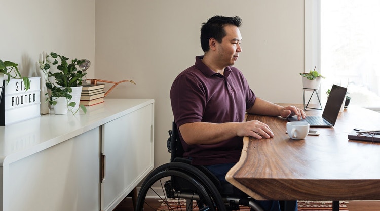 Man in a wheelchair and a maroon shirt writing a contract on his laptop at a wood table