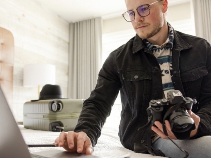 A photographer with camera in hand looking at a laptop while sitting on a bed next to a suitcase of equipment