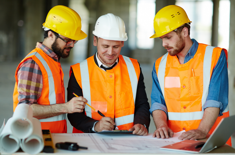 Three men wearing construction vests and safety helmets look at paperwork together
