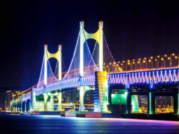 밤에 밝게 빛나는 현수교에는 다채로운 조명이 장식되어 있으며, 아래의 고요한 물 위로 반사됩니다.