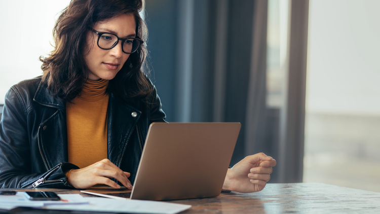 A woman sits at a table and looks at a laptop. She is wearing glasses and a leather jacket.
