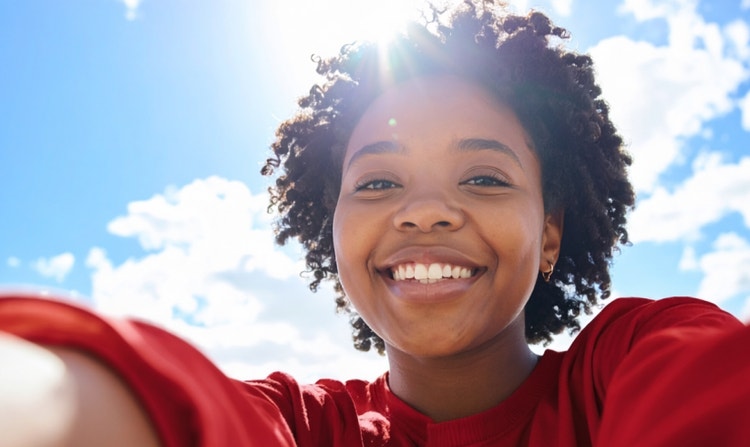 En smilende kvinne i rød skjorte tar en selfie utendørs i sollys mot en klar blå himmel, med detaljert hud og naturlig hårtekstur.