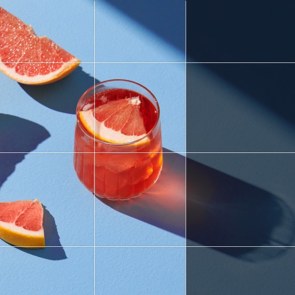 Isang larawan ng mga hiwa ng grapefruit sa tabi ng pink na cocktail na pinalaki para makita ang buong anino ng cocktail glass.