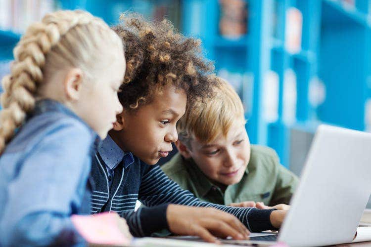 Grupo de tres diferentes estudiantes que demuestran entusiasmo mientras usan una computadora portátil y están en el escritorio de la biblioteca escolar
