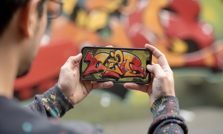 A person holds a smartphone showing colorful graffiti artwork, with a blurred graffiti mural in the background.
