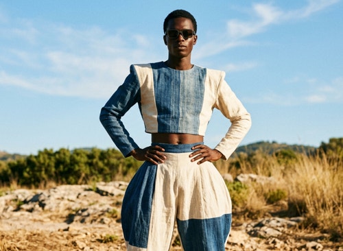 Model wearing sunglasses and a sculptural blue-and-white outfit stands outdoors against a clear sky and natural landscape.