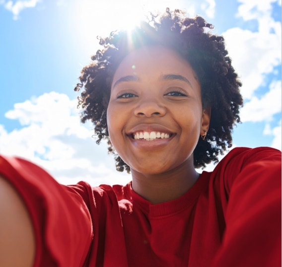 Smiling woman in a red shirt takes a sunlit outdoor selfie against a bright blue sky, with detailed skin and natural hair texture.