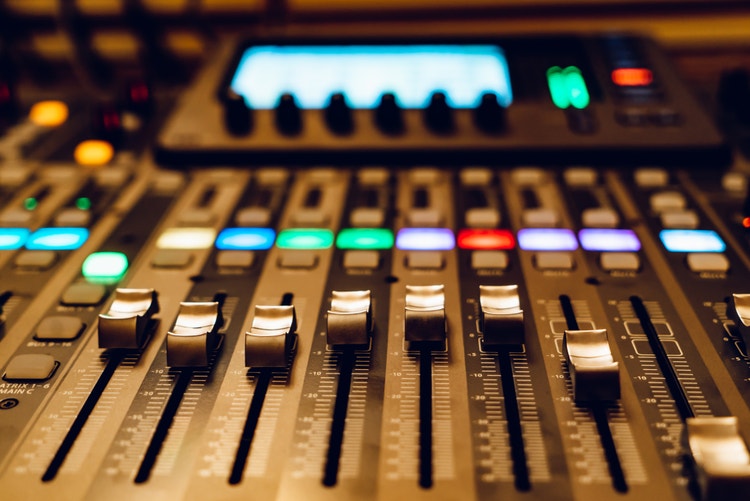 Close-up view of an audio mixing console with multiple faders and knobs, illuminated by colorful LED lights.