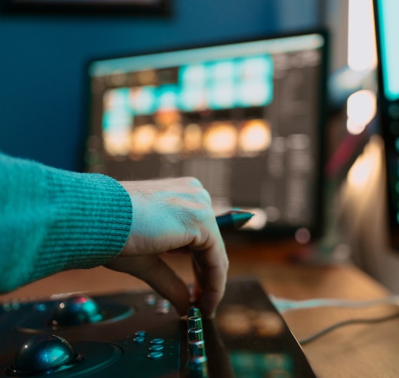 A person using an audio editing console beside computer monitors displaying an editing timeline. The workspace illustrates integrating AI-generated voiceovers into a broader production workflow from recording through final editing.