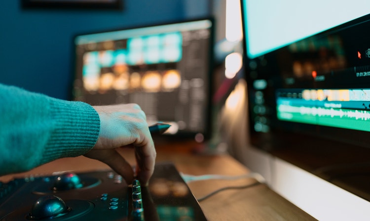 A person using an audio editing console beside computer monitors displaying an editing timeline. The workspace illustrates integrating AI-generated voiceovers into a broader production workflow from recording through final editing.
